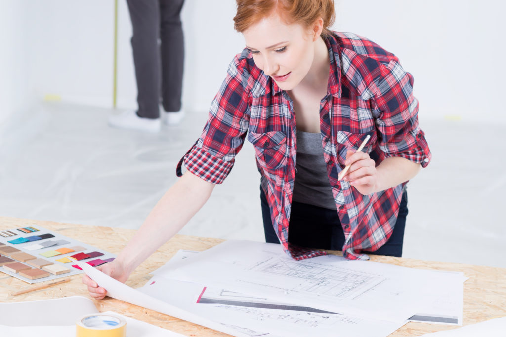 Young and red haired woman designer leaning on desk with interior projects and color picker, managing design budget.